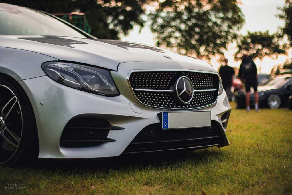 fahrzeugfotografie_ein_silberner_mercedes_steht_auf_einer_gruenen_wiese_im_hintergrund_ein_paar_baeume_und_zwei_personen