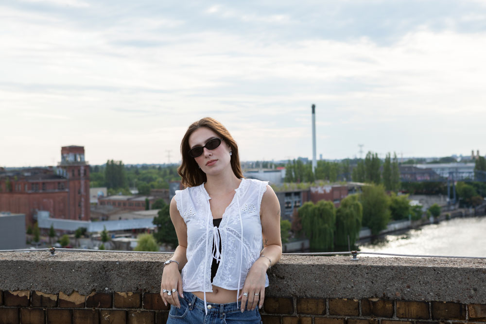 eine_frau_mit_sonnenbrille_lehnt_laessig_an_einer_wand_im_hintergrund_die_spree_und_ein_industriegebiet